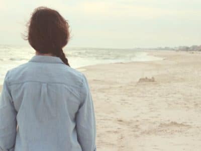 Young woman with plait hair with her back to you standing on the beach looking at the sand and sea