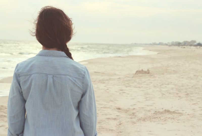 Young woman with plait hair with her back to you standing on the beach looking at the sand and sea