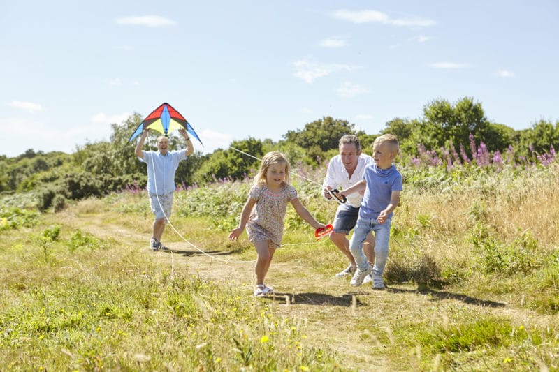 Adoptive male parents running with a kite and playing with their primary school age children on a Devon path, on a sunny day