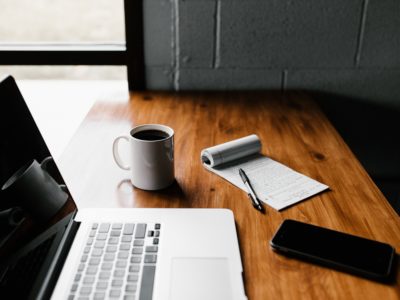 A desk with an open laptop, a mobile phone, a notepad and pen and mug of coffee