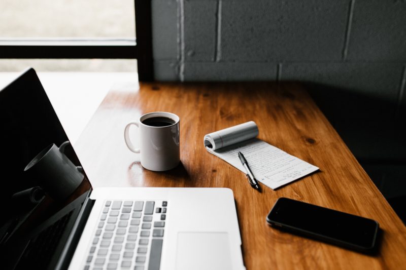 A desk with an open laptop, a mobile phone, a notepad and pen and mug of coffee