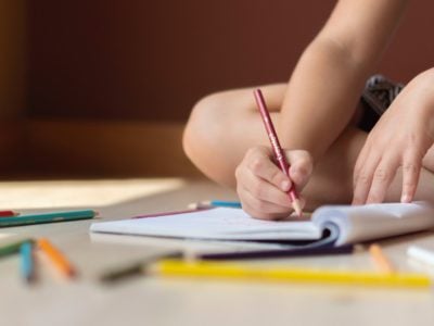 a person writing in book surrounded by pencil crayons