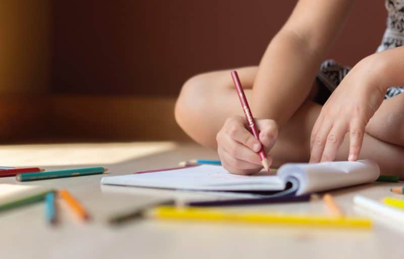 a person writing in book surrounded by pencil crayons